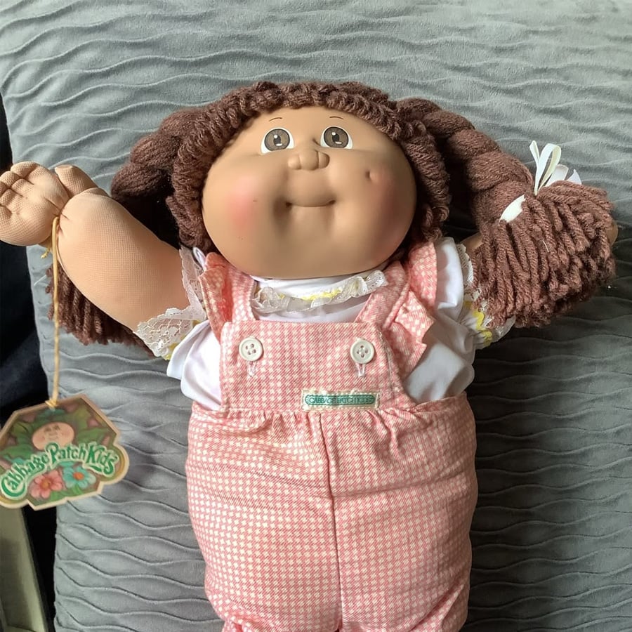 A Cabbage Patch Kids doll with brown yarn hair, wearing pink gingham overalls and a white shirt, lies on a gray textured surface. The doll has a smiling face with dimples and holds a tag in its left hand.