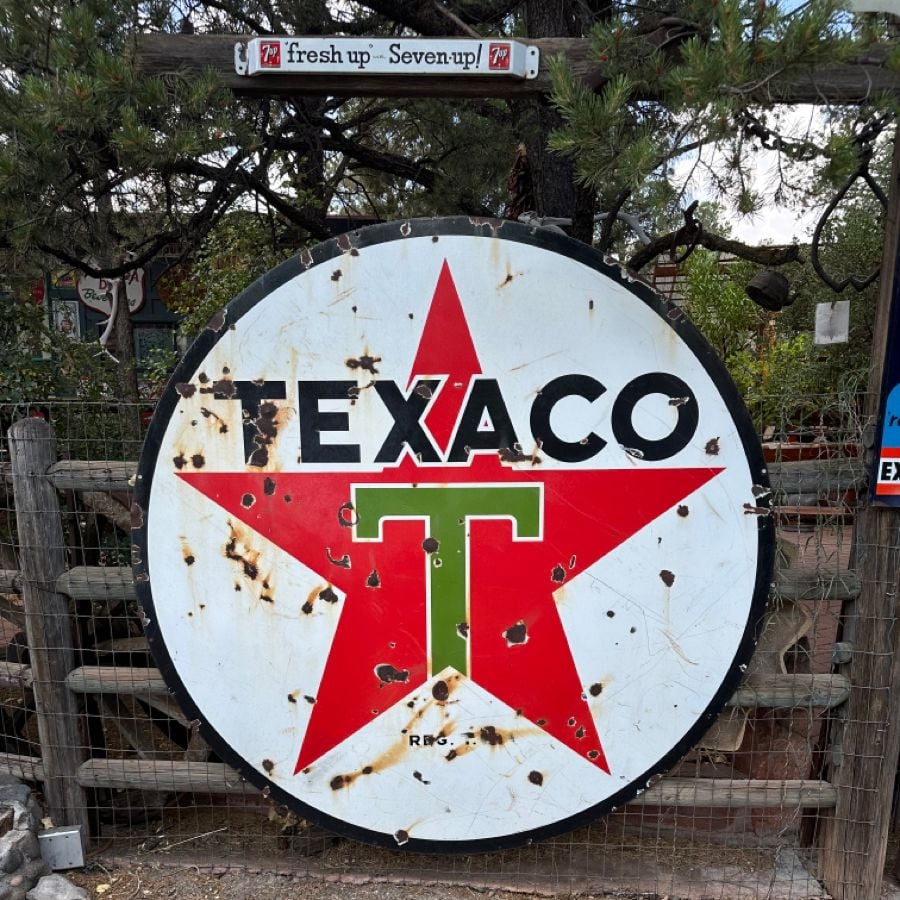 A large, weathered Texaco sign with a red star and green "T" stands in front of a wooden fence. There is a small "fresh up - Seven-Up!" sign above it, surrounded by trees and foliage.