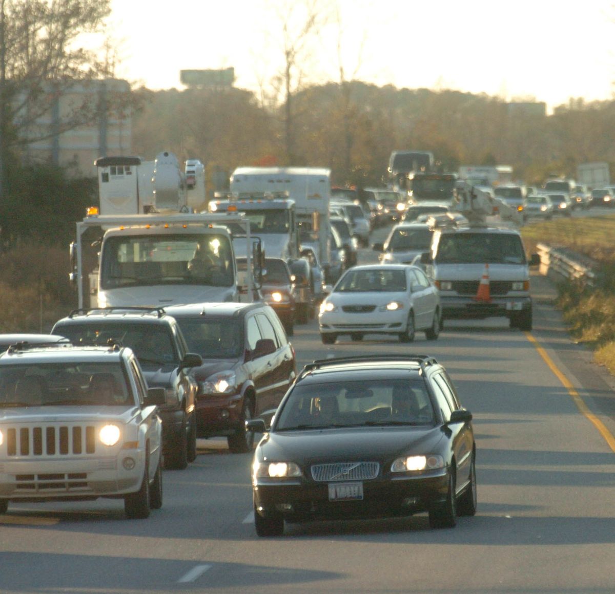 Heavy traffic with cars and trucks fills multiple lanes on a highway during daylight, with some vehicles stopped and others moving slowly. Trees and a barrier line the road.