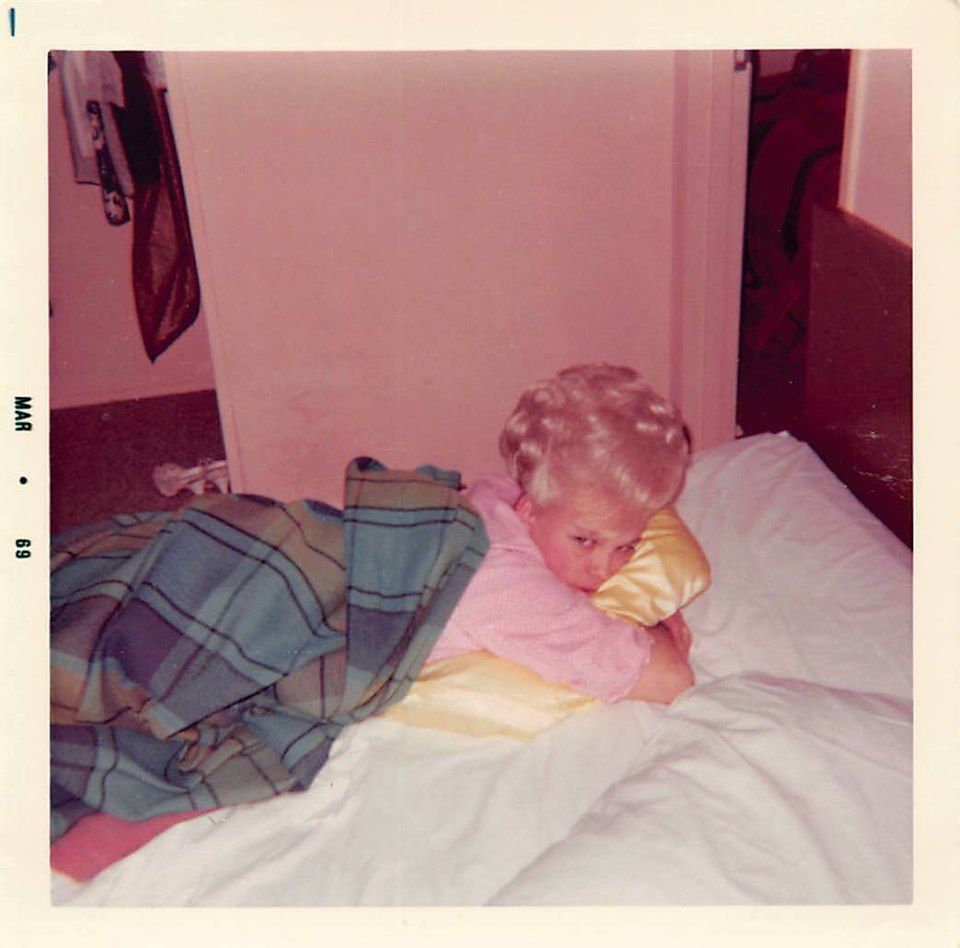 A young child with blonde hair lies on a bed, resting on a yellow pillow and covered partly with a plaid blanket. The photo has a vintage look and is dated "May 69" along the edge.