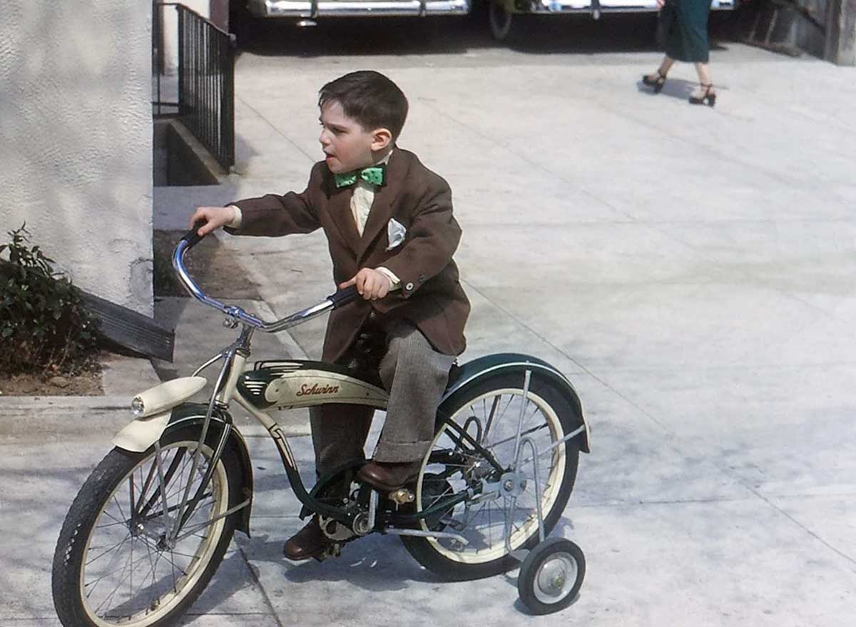 A young boy in a brown suit and green bow tie rides a tricycle labeled "Schwinn" on a sidewalk, looking to his right. An adult walks by in the background.