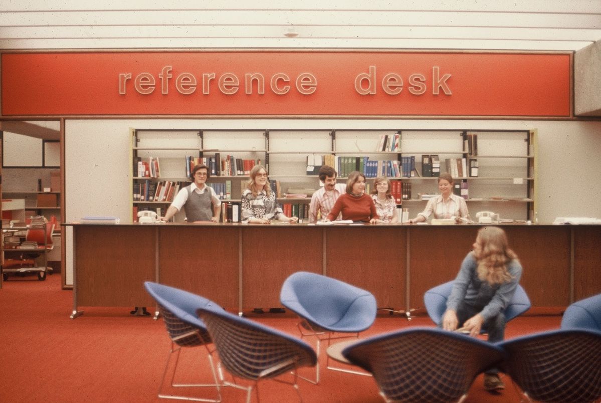 Six people stand behind a library reference desk with bookshelves behind them, and one person sits in a blue chair in the foreground. The floor and walls are red, and a large "reference desk" sign is displayed above.