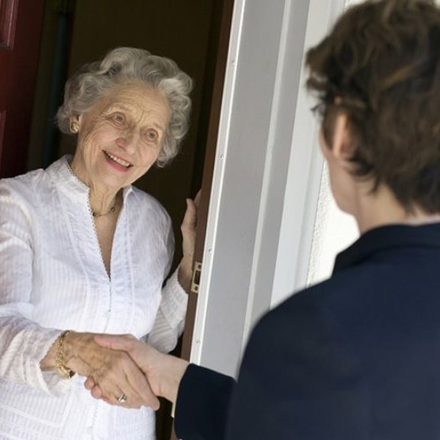 An elderly woman in a white blouse smiles while shaking hands with a younger person at her front door.