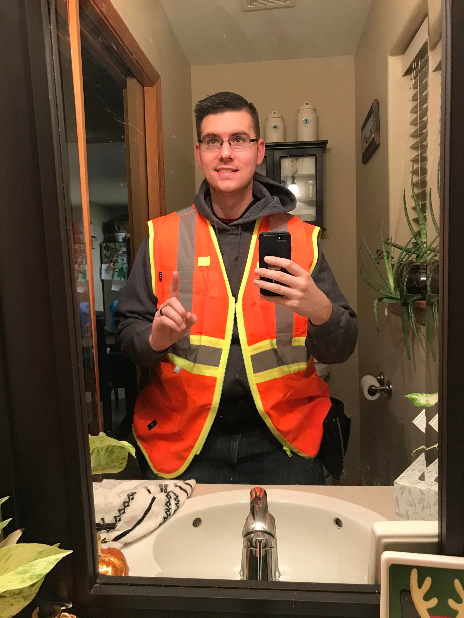 A person wearing an orange safety vest and gray hoodie takes a mirror selfie in a bathroom, holding a phone and making a peace sign with their hand.