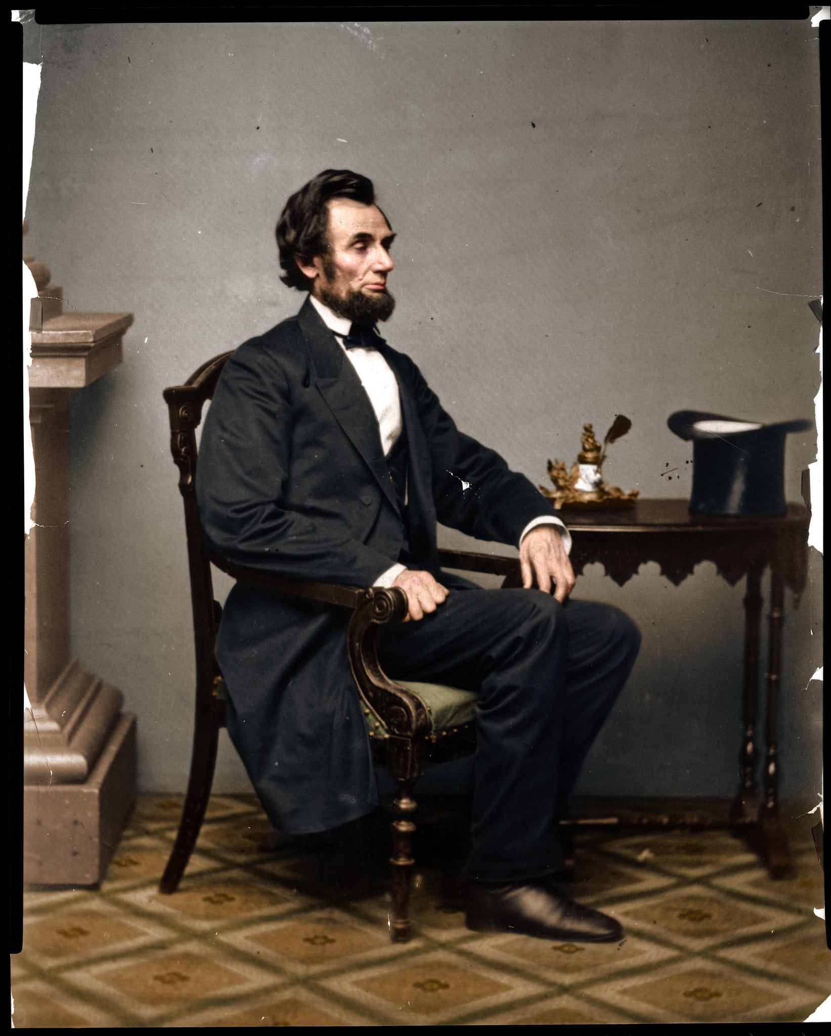 A man in a dark suit sits sideways in an ornate chair beside a small table with a hat, papers, and a quill, against a plain background.