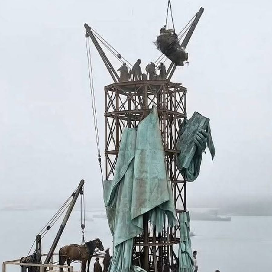 Workers assemble a large metal statue on scaffolding, draping copper sheets over the structure. A crane lifts the statue's head, while a horse and more workers stand below. The background is foggy, with water visible in the distance.