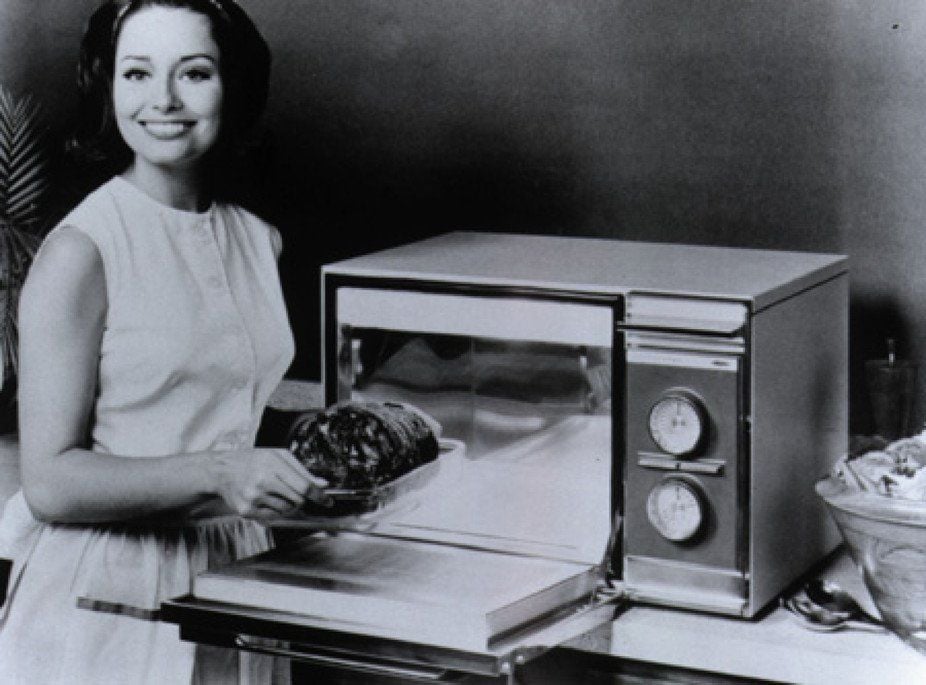 A woman in a dress smiles while placing a roast into a vintage microwave oven with dials, on a kitchen counter. Black and white photo.