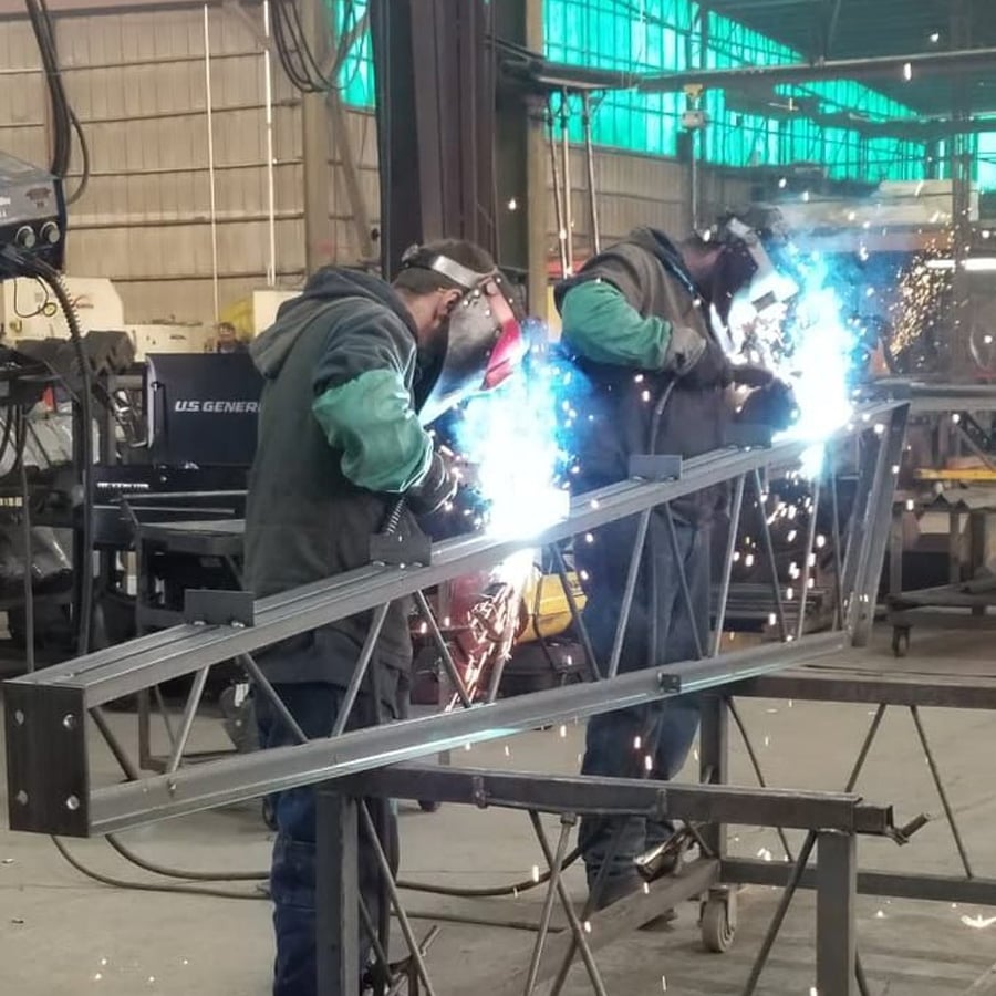 Two people wearing protective gear are welding metal beams in an industrial workshop. Bright sparks and blue light from welding torches are visible, with equipment and machinery in the background.