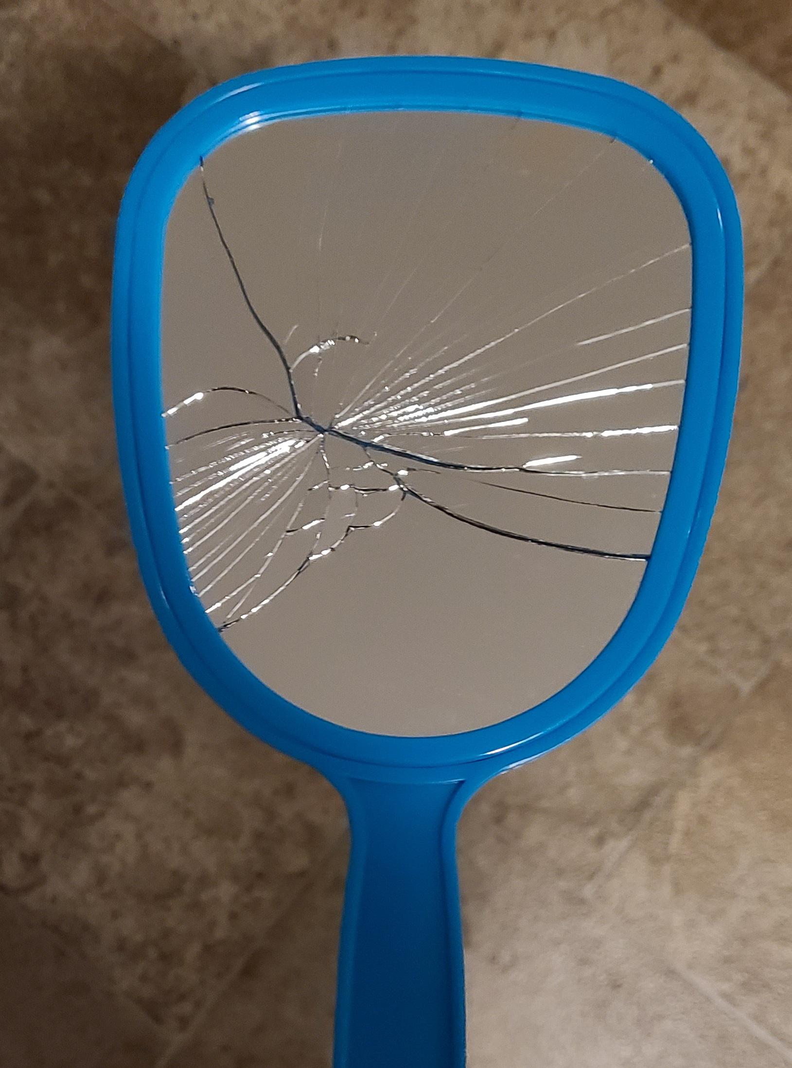 A blue handheld mirror with a cracked and shattered reflective surface lies on a brown textured surface.