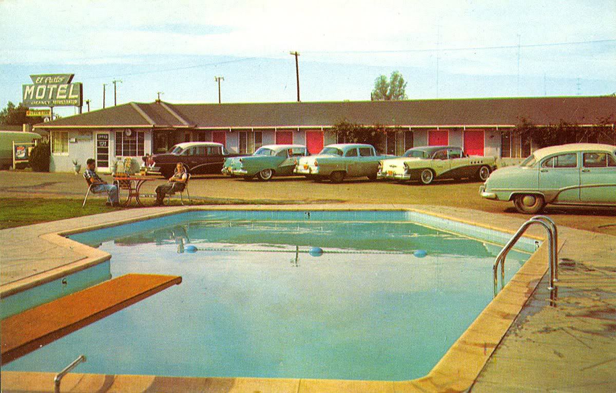 A vintage motel scene with classic cars parked outside, people sitting by a pool, and a neon motel sign in the background. The pool has a diving board and clear blue water.