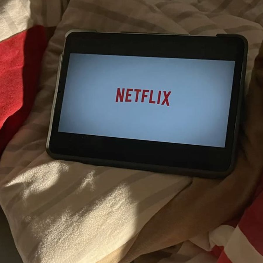 A tablet displaying the Netflix logo rests on a bed with striped and solid-colored blankets in dim lighting.