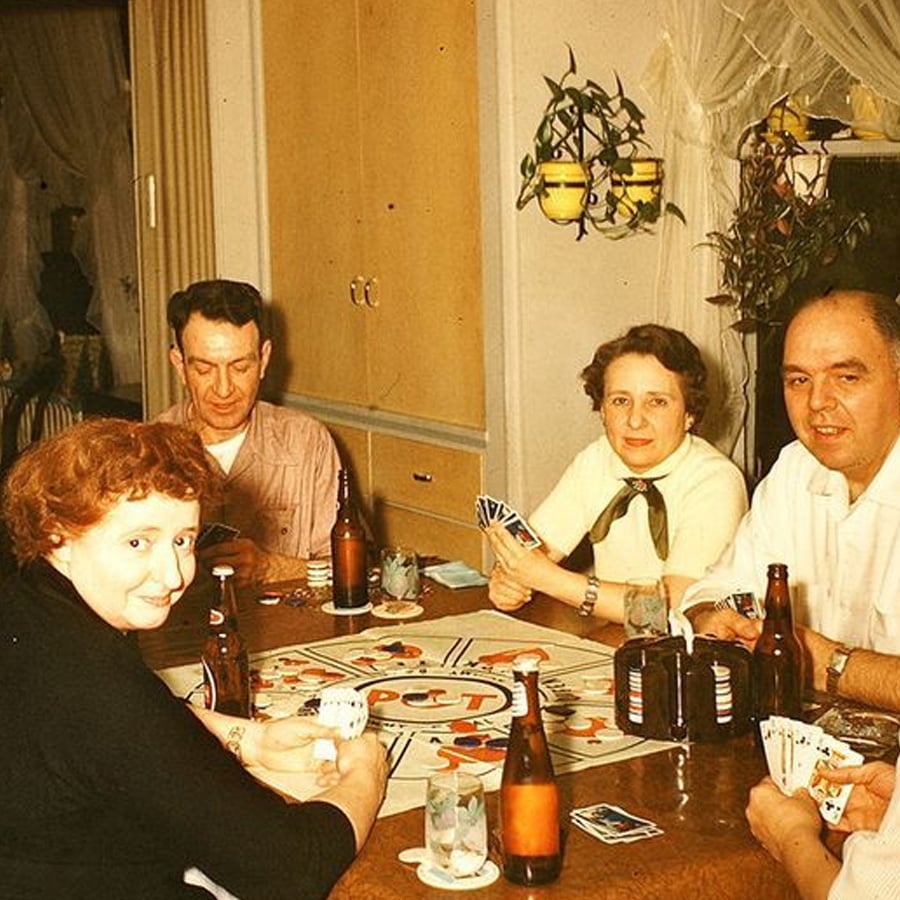 Four adults sit around a table playing cards and a board game, drinks on the table, with plants and a cabinet in the background. Everyone appears relaxed and engaged in the game.
