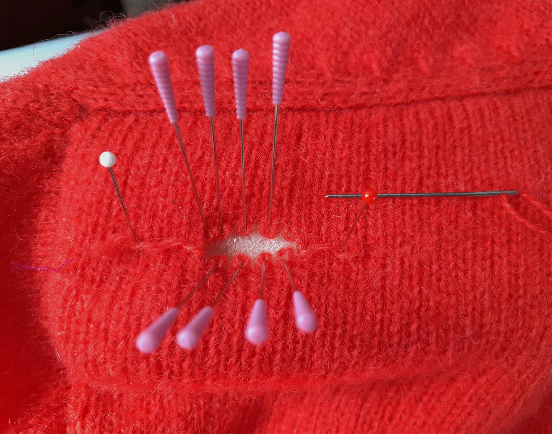 A red knitted fabric with a hole being mended. Several pink and red pins hold the fabric in place while a needle with red thread is being used to sew around the damaged area.