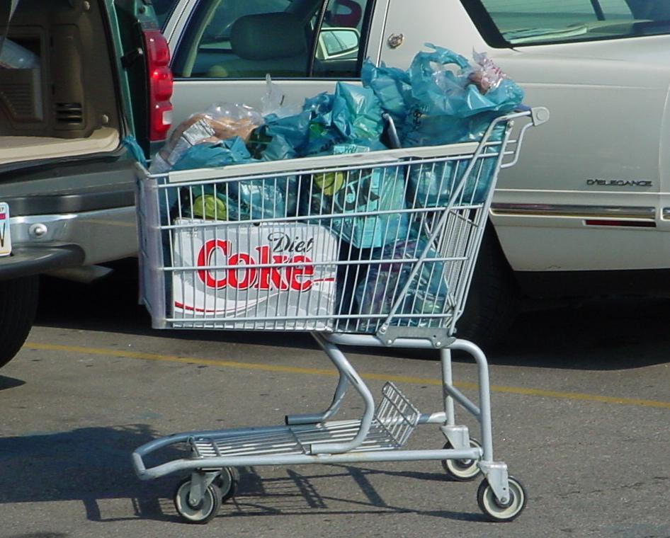 A shopping cart filled with groceries in blue plastic bags sits in a parking lot next to a white car and an open SUV trunk. The cart displays a Diet Coke advertisement on its side.