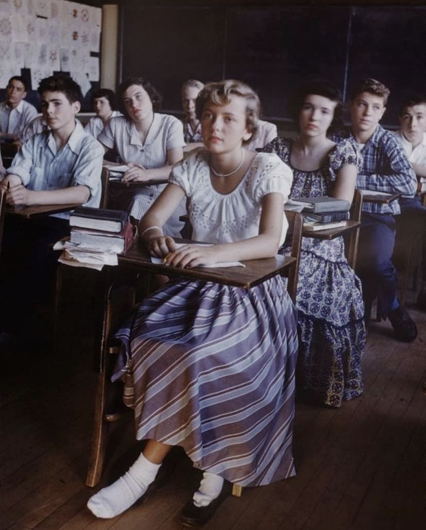 A group of students sit at wooden desks in a classroom, facing forward. Most wear 1950s-style clothing. The girl in front has one shoe off, resting her socked foot on a rung. Books and papers cover the desks.