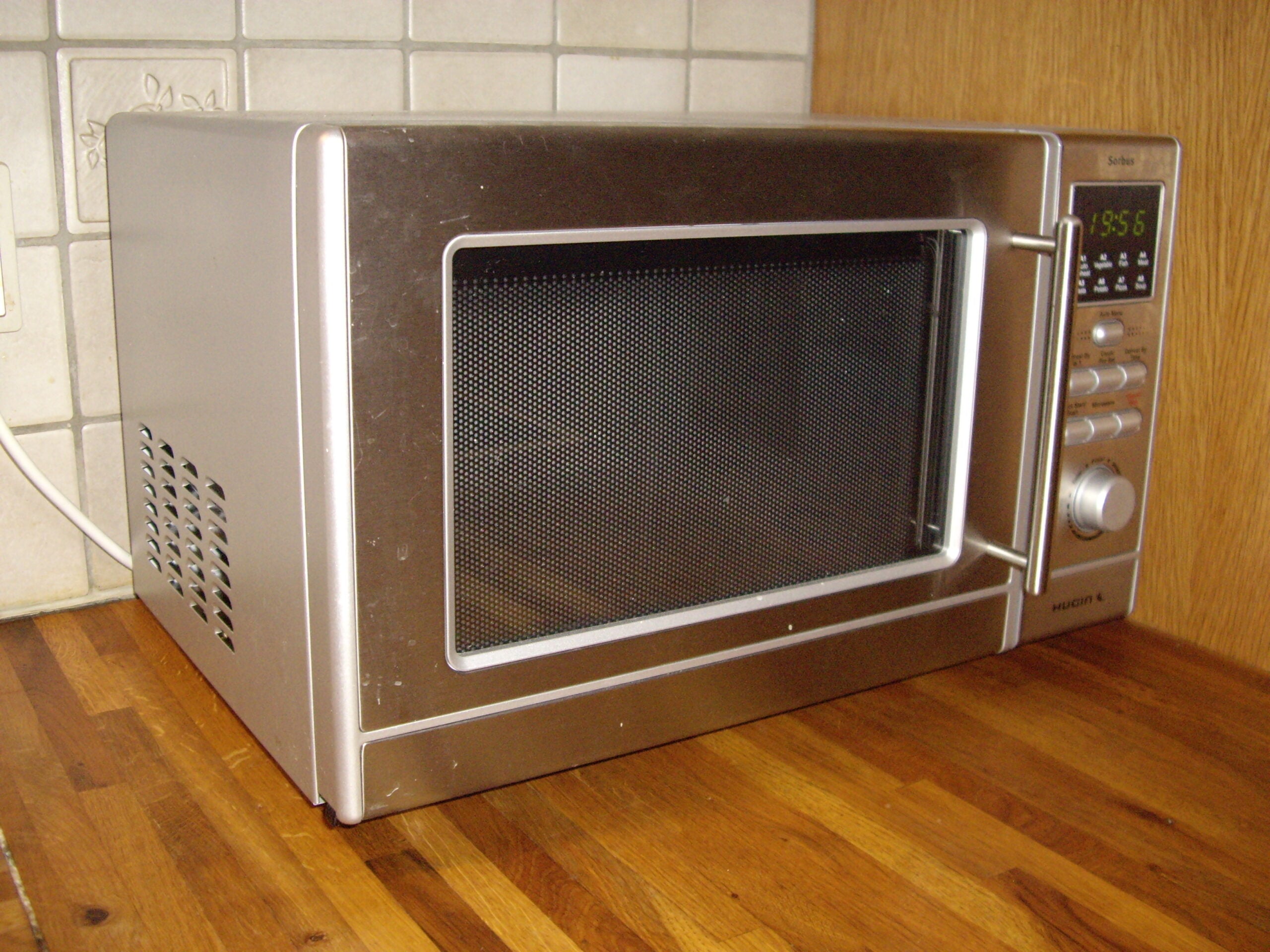 A stainless steel microwave oven sits on a wooden kitchen countertop, with a digital display, control buttons, and a handle on the right side. Tile backsplash and a wood wall are visible behind it.