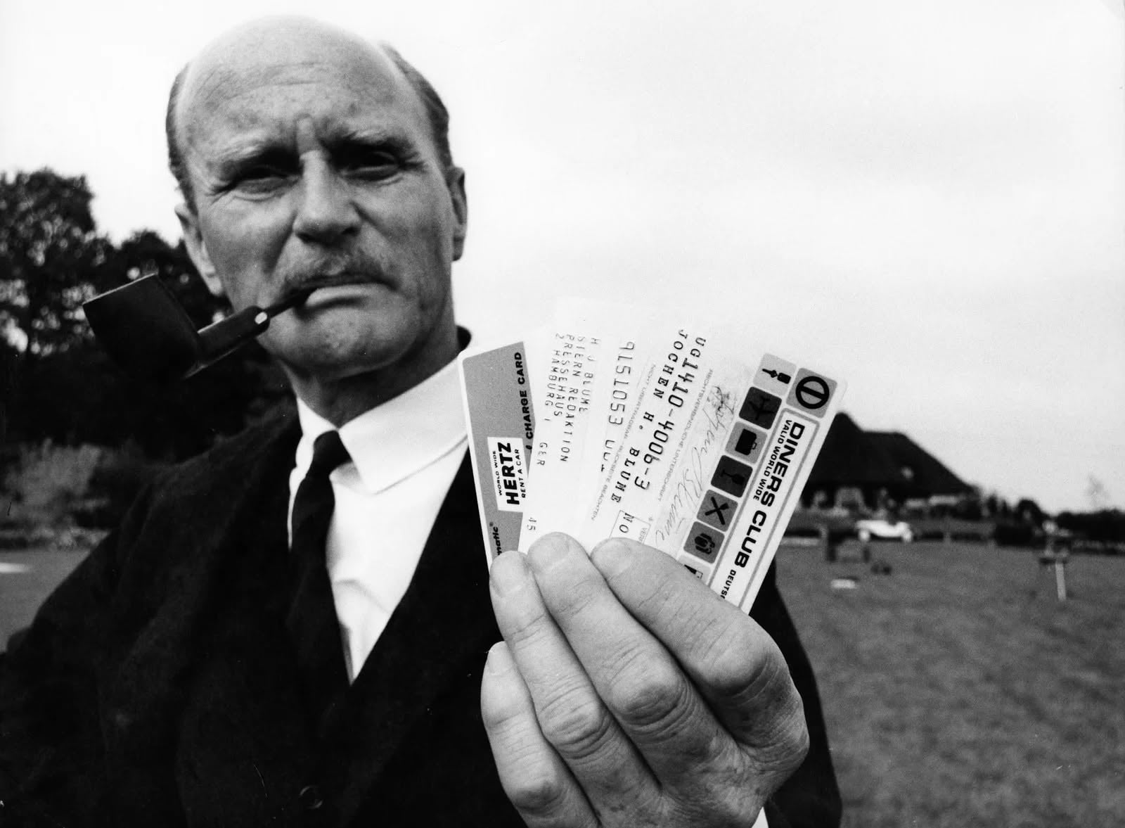 A man in a suit with a pipe in his mouth holds up a Diners Club card and two tickets outdoors, with a grassy field and buildings visible in the background.