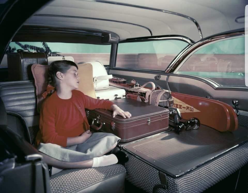 A child in a red sweater sleeps in the backseat of a car, surrounded by luggage and a violin case on the back shelf. The interior looks spacious and vintage.