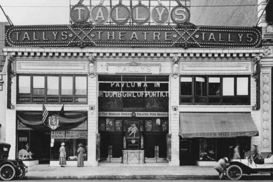 Black and white photo of Tally’s Theatre with a marquee sign, people standing outside, vintage cars parked along the street, and shops on either side of the theater entrance.