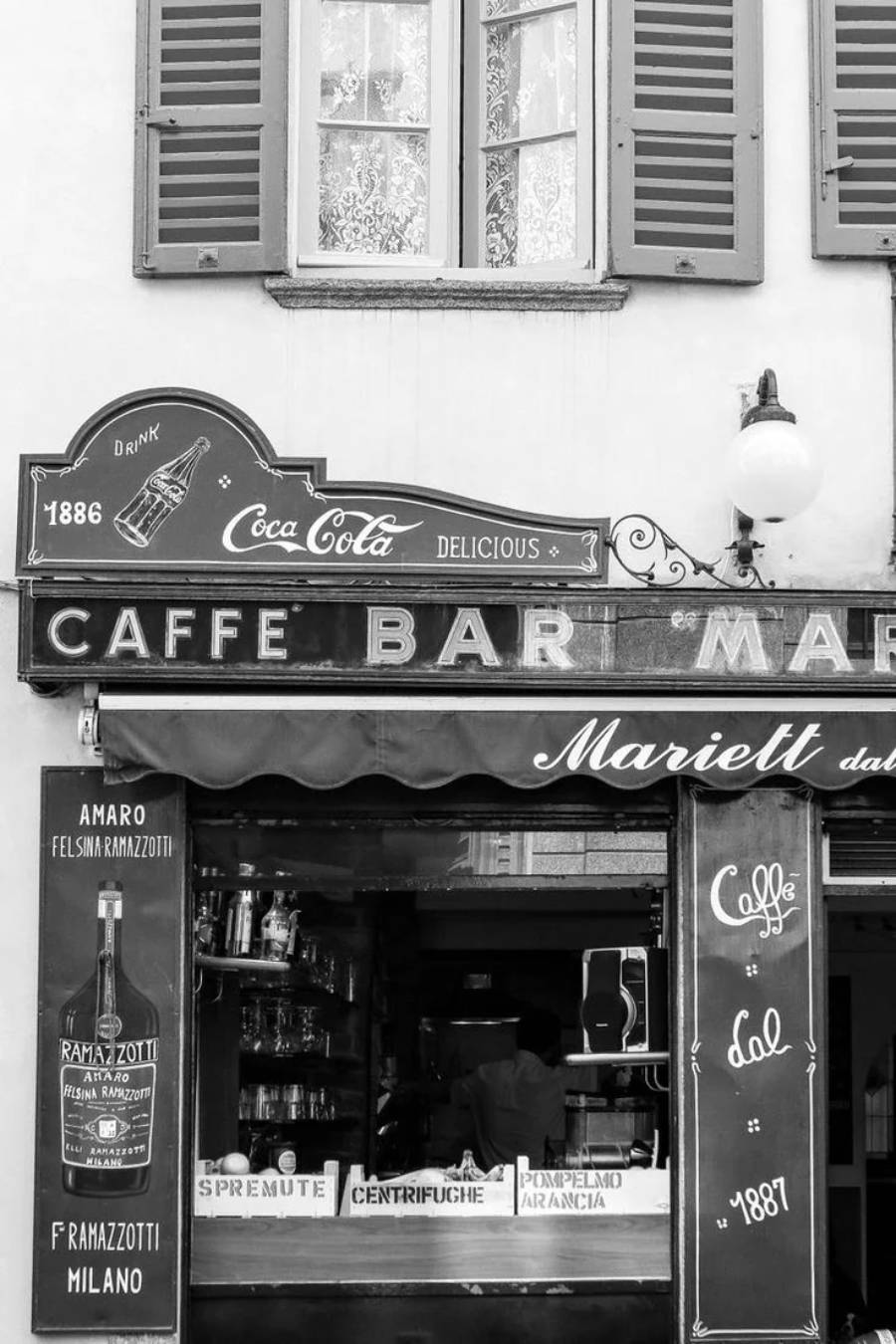 Black and white photo of a vintage Italian café bar with Coca-Cola and Ramazzotti signs, an awning, open window counter displaying bottles, and shuttered windows above.