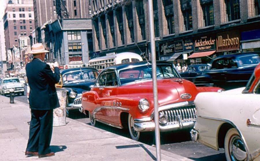 A man in a suit and hat stands on a city sidewalk next to a row of vintage cars, including a bright red Buick, with tall buildings and busy street traffic in the background.