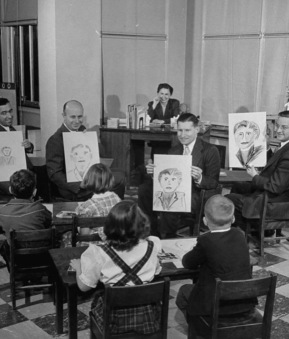 Four men seated in front of a group of schoolchildren each hold up a portrait drawing. A teacher sits at a desk in the background, observing the scene in a classroom setting.