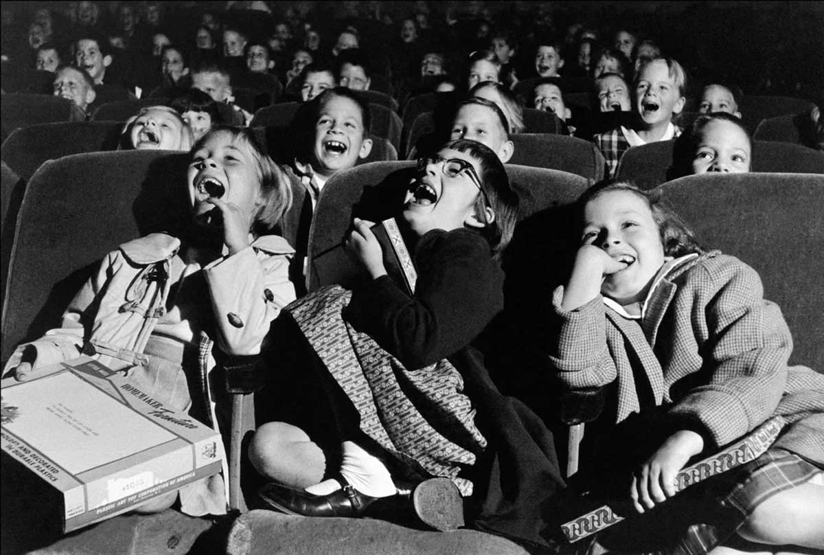 A group of children sit in theater seats, laughing and smiling joyfully as they watch a show or movie. Several hold candy or snack boxes, and the atmosphere is lively and cheerful.