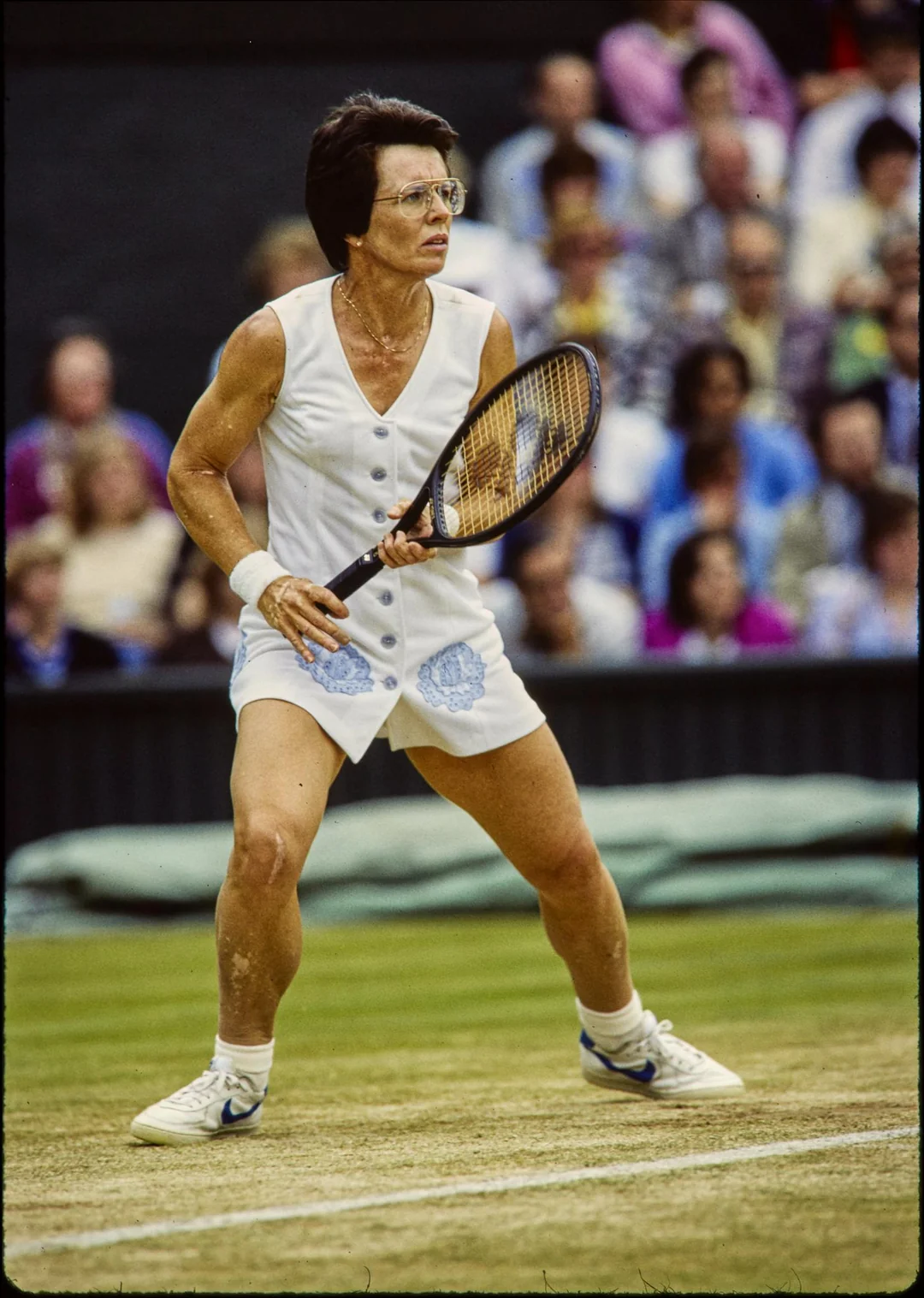 A female tennis player in a white outfit holds a racquet and prepares to return a shot on a grass court, with spectators watching in the background.