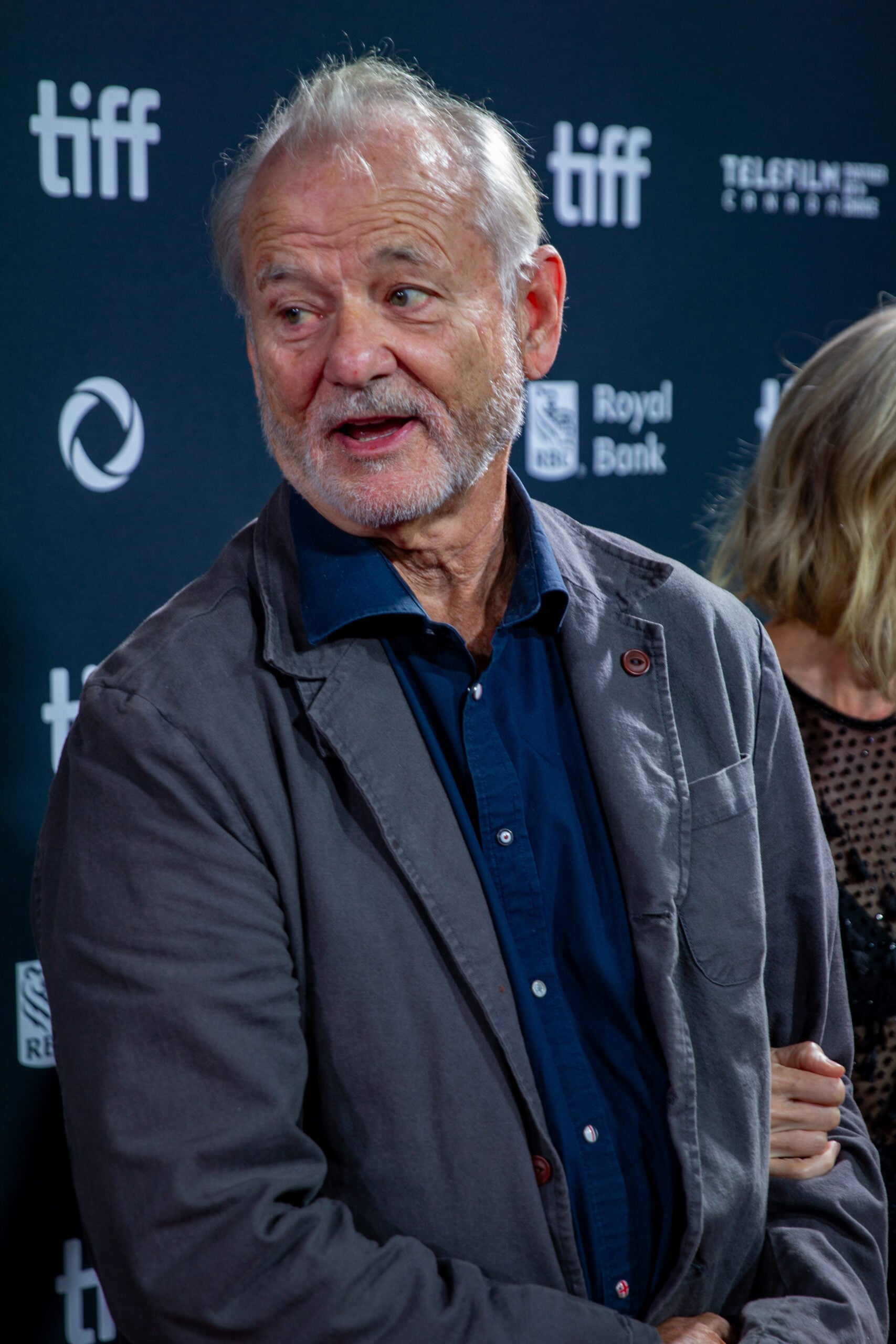 An older man with gray hair and a beard, wearing a blue shirt and gray jacket, stands in front of a backdrop with "tiff" and sponsor logos at an event.