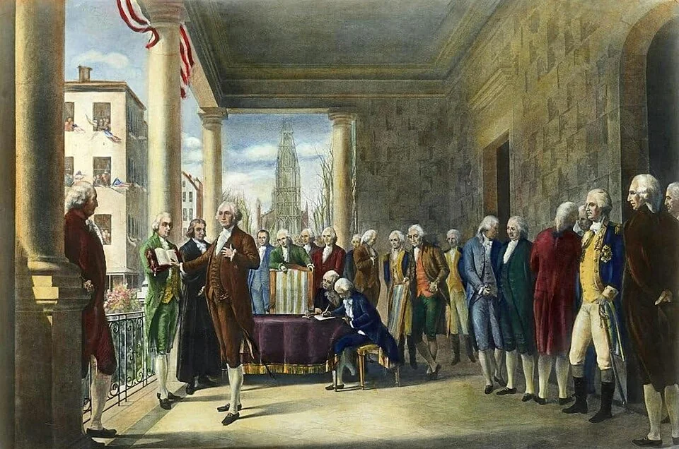 A group of men in 18th-century clothing stand on a balcony, with one man placing his hand on a book while others watch; an American flag is above them.