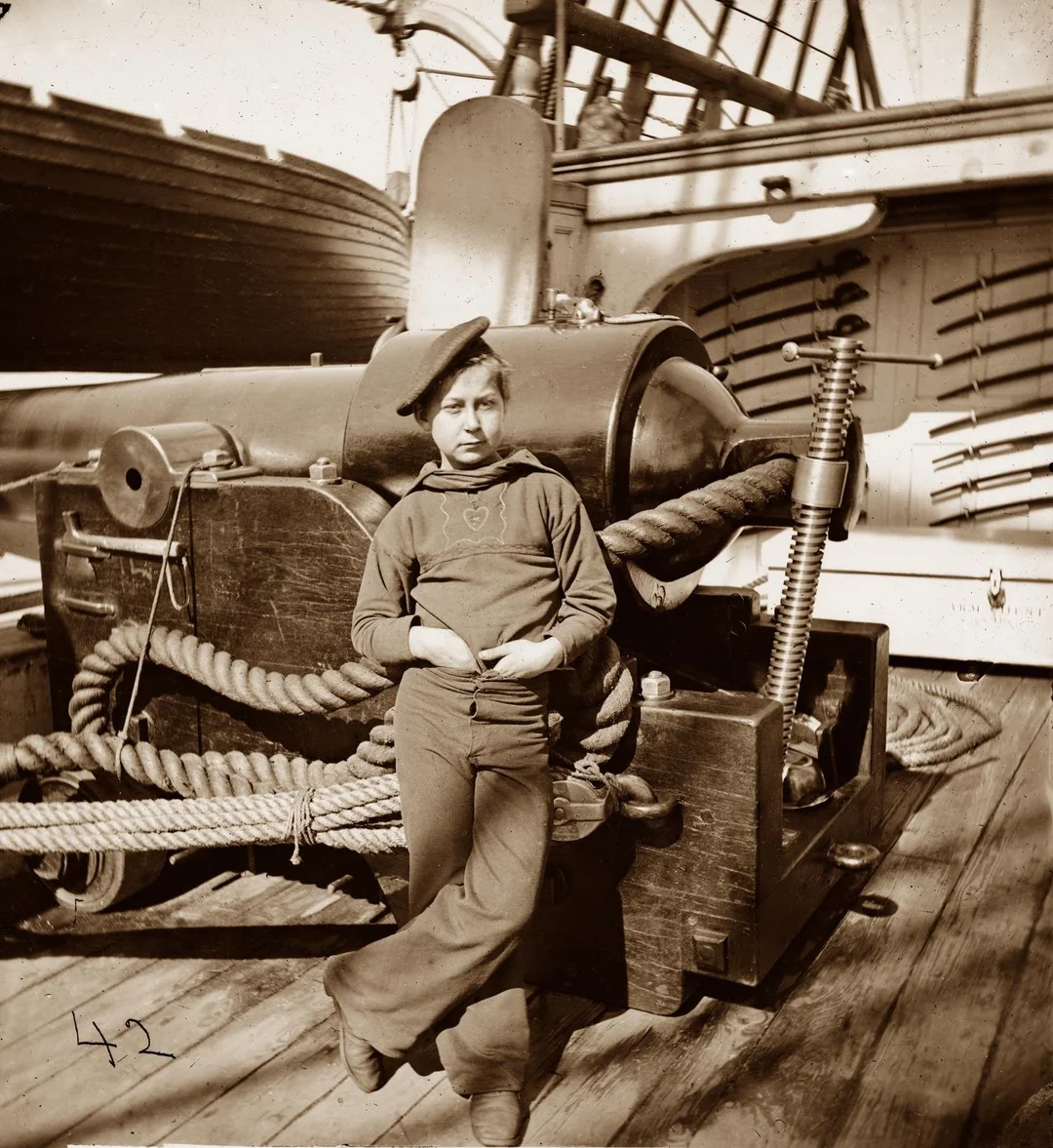 Young person in sailor uniform leans against ship machinery on a wooden deck, with coiled ropes and rifles visible in the background, aboard a historical ship.