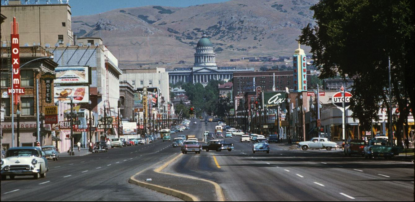 A busy city street from the 1950s or 1960s with vintage cars, neon signs, shops, and a large domed capitol building in the background against mountain hills.
