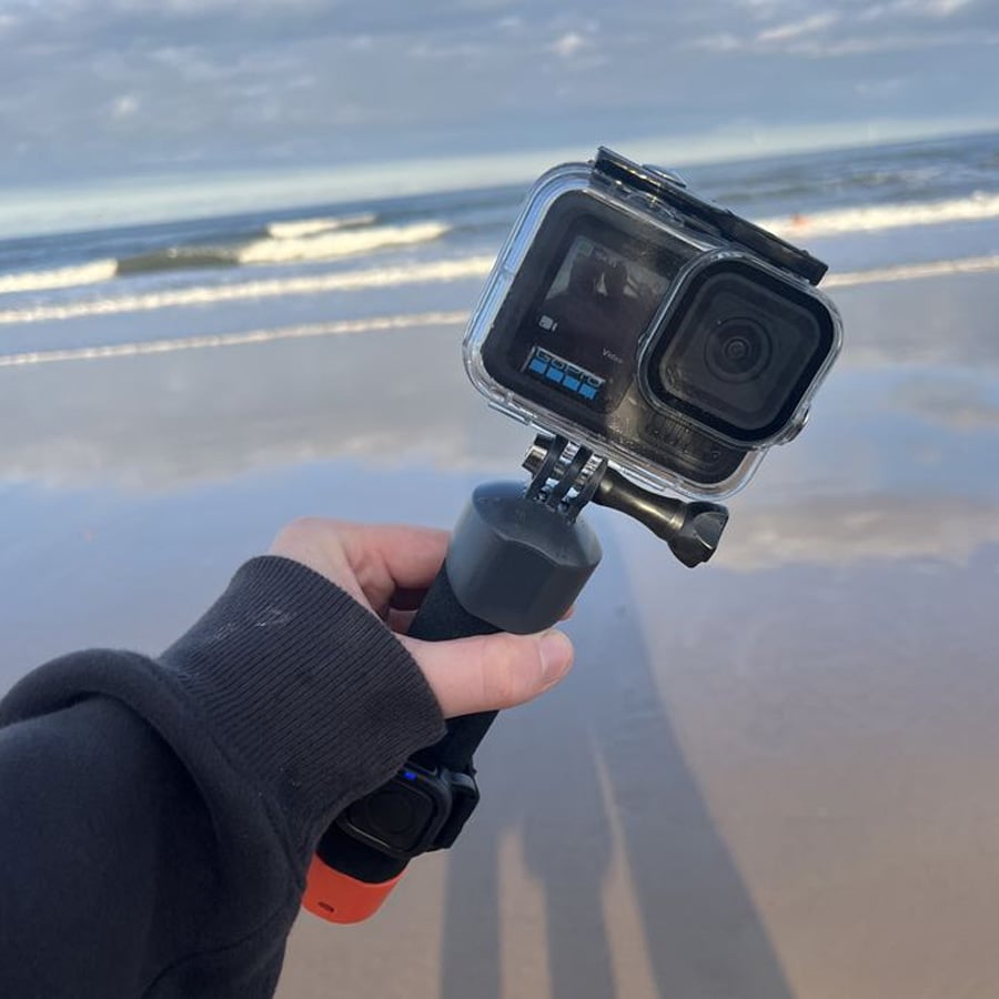 A person holding a GoPro camera with a grip is standing on a sandy beach, with waves and a cloudy sky visible in the background.