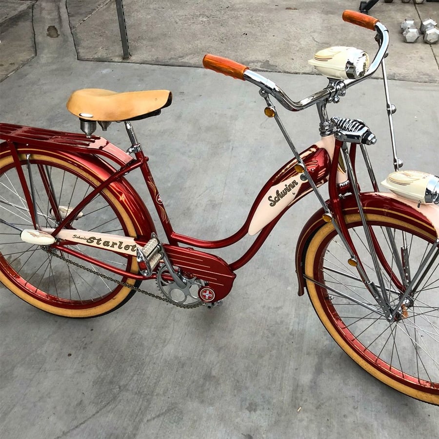 A vintage red and cream Schwinn bicycle with tan tires, chrome details, and a retro headlight, parked on a concrete surface.