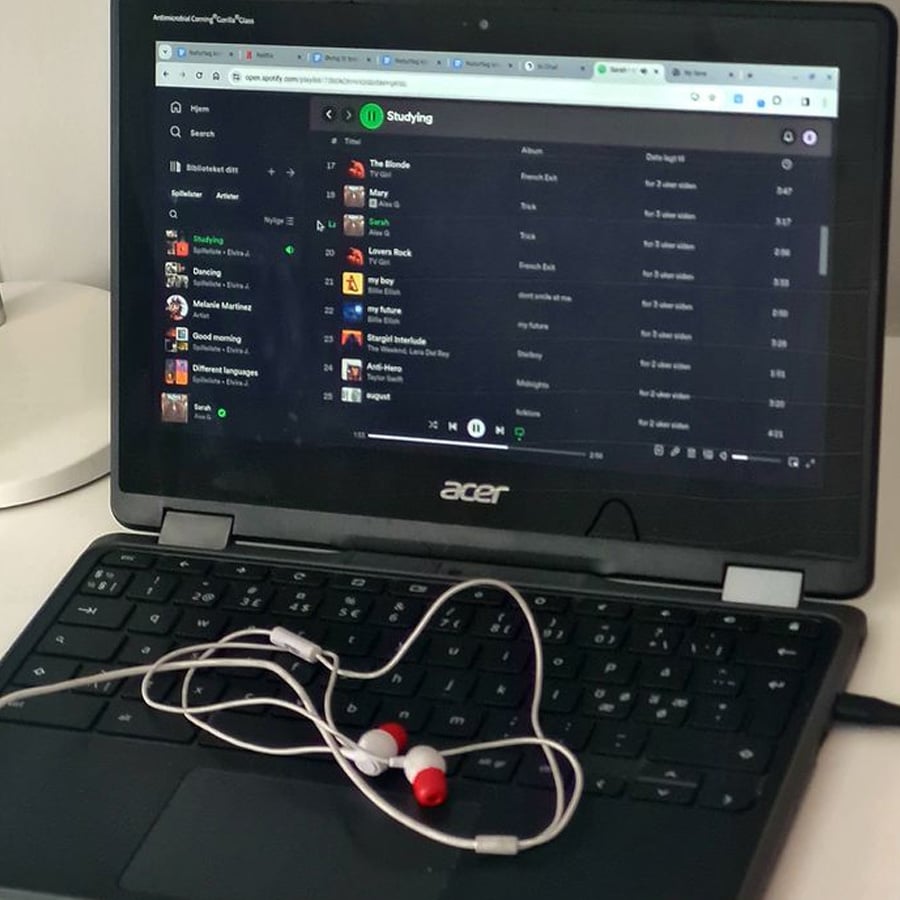 A black Acer laptop displays a Spotify playlist called “Studying.” A pair of white and red wired earphones rests on the keyboard.