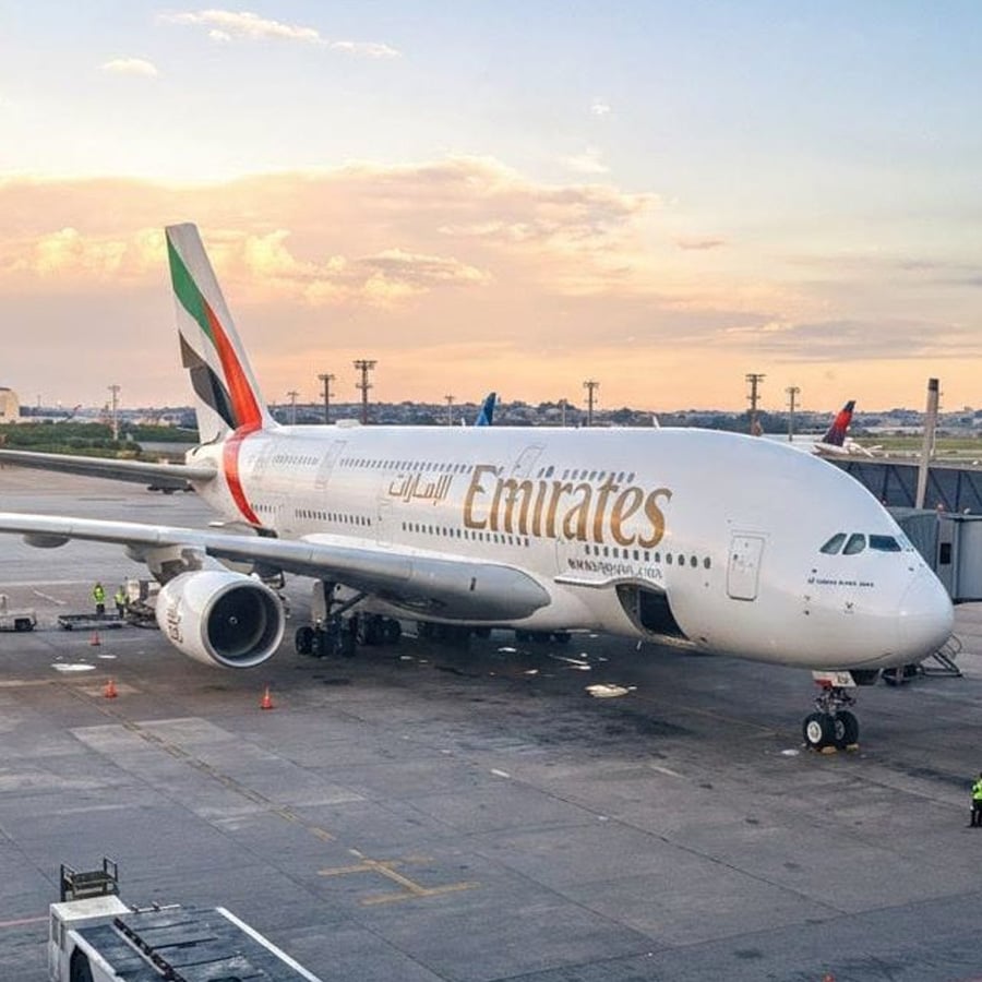 An Emirates Airbus A380 is parked at an airport gate during sunset, with ground crew and equipment nearby. The sky is partly cloudy with orange and blue hues.