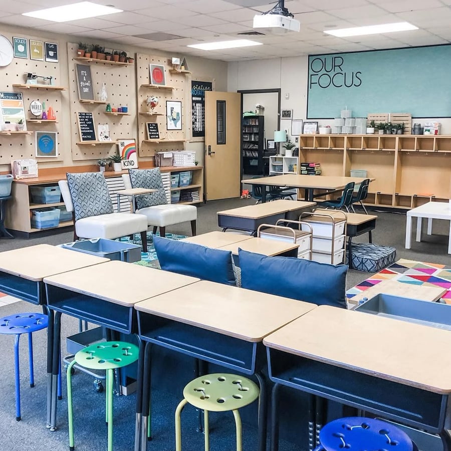 A modern classroom with desks, colorful stools, couches, shelves, pegboards with decorations, and a wall displaying "OUR FOCUS." The room has a cozy, organized, and flexible seating arrangement.