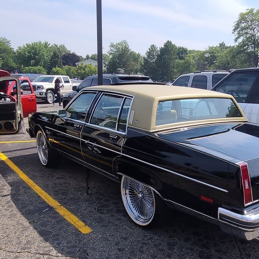 A black classic car with a tan vinyl roof is parked in a lot among other vehicles on a sunny day. The car has shiny chrome rims and tinted windows. Trees are visible in the background.