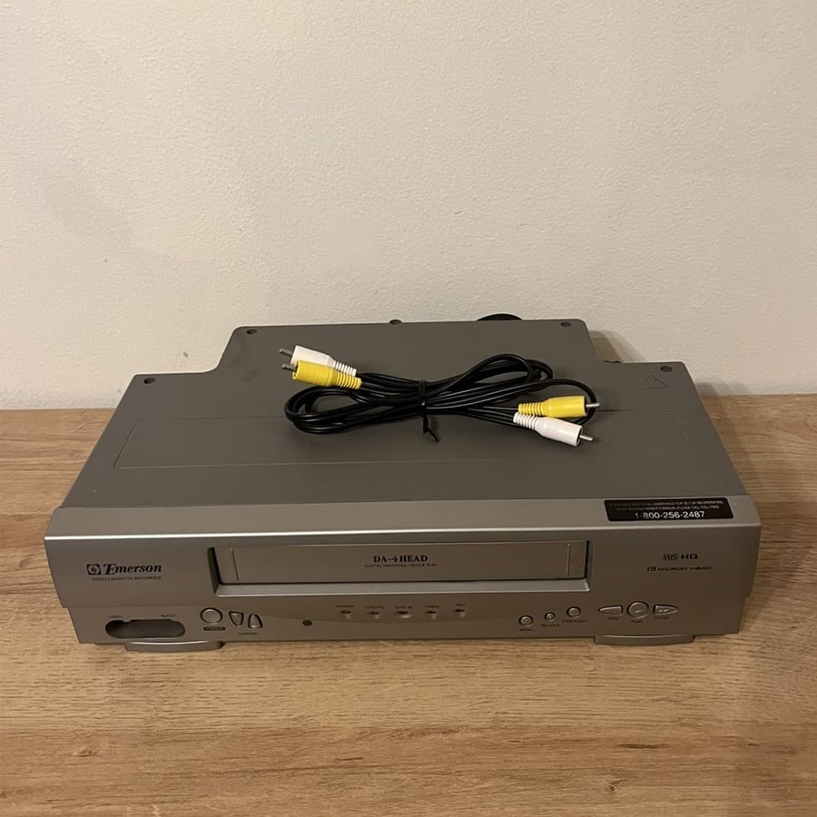 A silver Emerson VCR sits on a wooden surface with a black, yellow, and white RCA cable coiled on top.