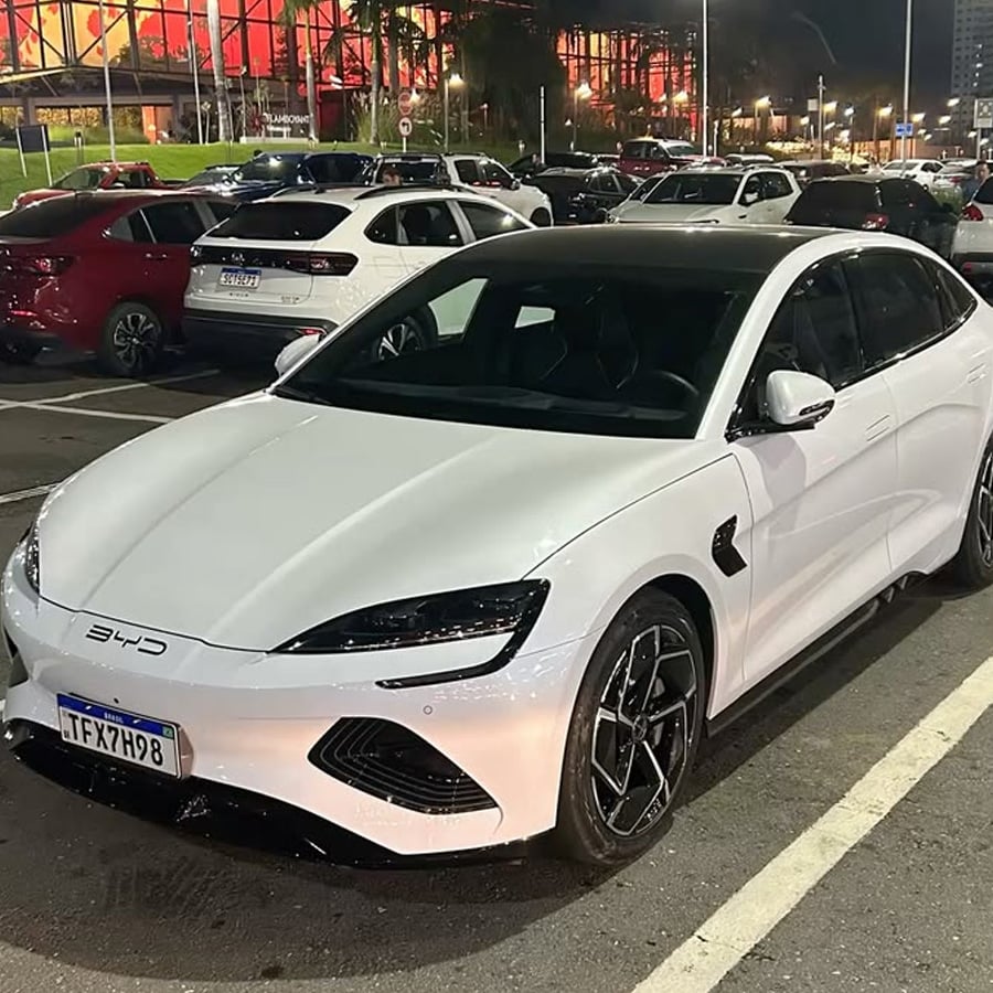 A white BYD electric car is parked in a well-lit parking lot at night, surrounded by other vehicles. Buildings and streetlights are visible in the background.
