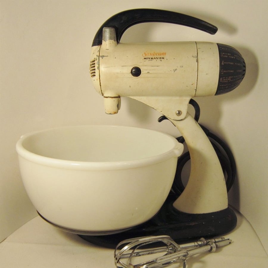 Vintage Sunbeam Mixmaster stand mixer with a white mixing bowl. The mixer is off-white with black accents, and a pair of metal beaters is placed in front of the bowl. The mixer shows signs of wear.
