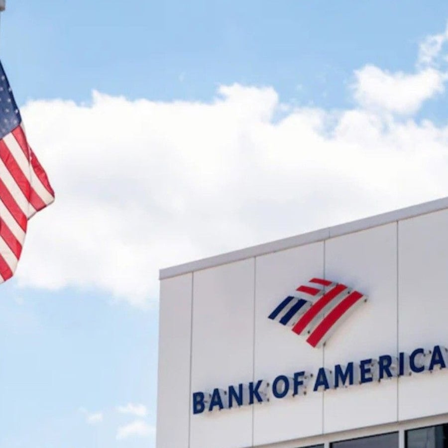 A U.S. flag waves near the corner of a Bank of America building, with the bank's logo and name visible against a partly cloudy sky.