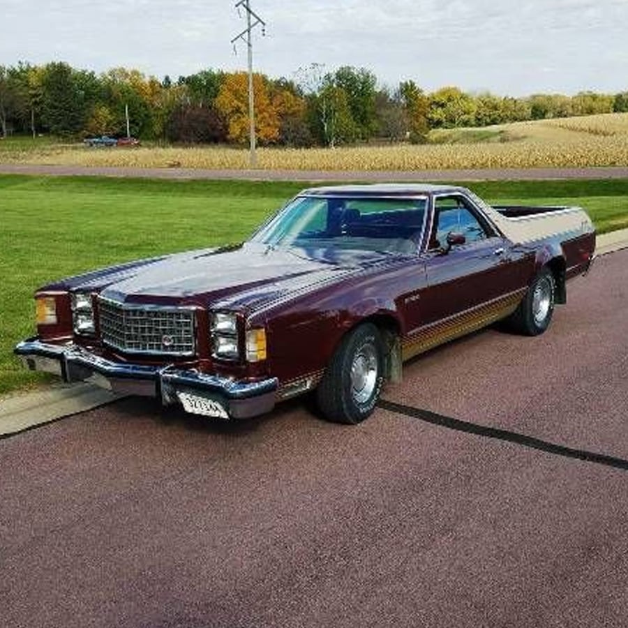 A vintage maroon Ford Ranchero is parked on a paved road beside a grassy field with trees and crops in the background under a partly cloudy sky.