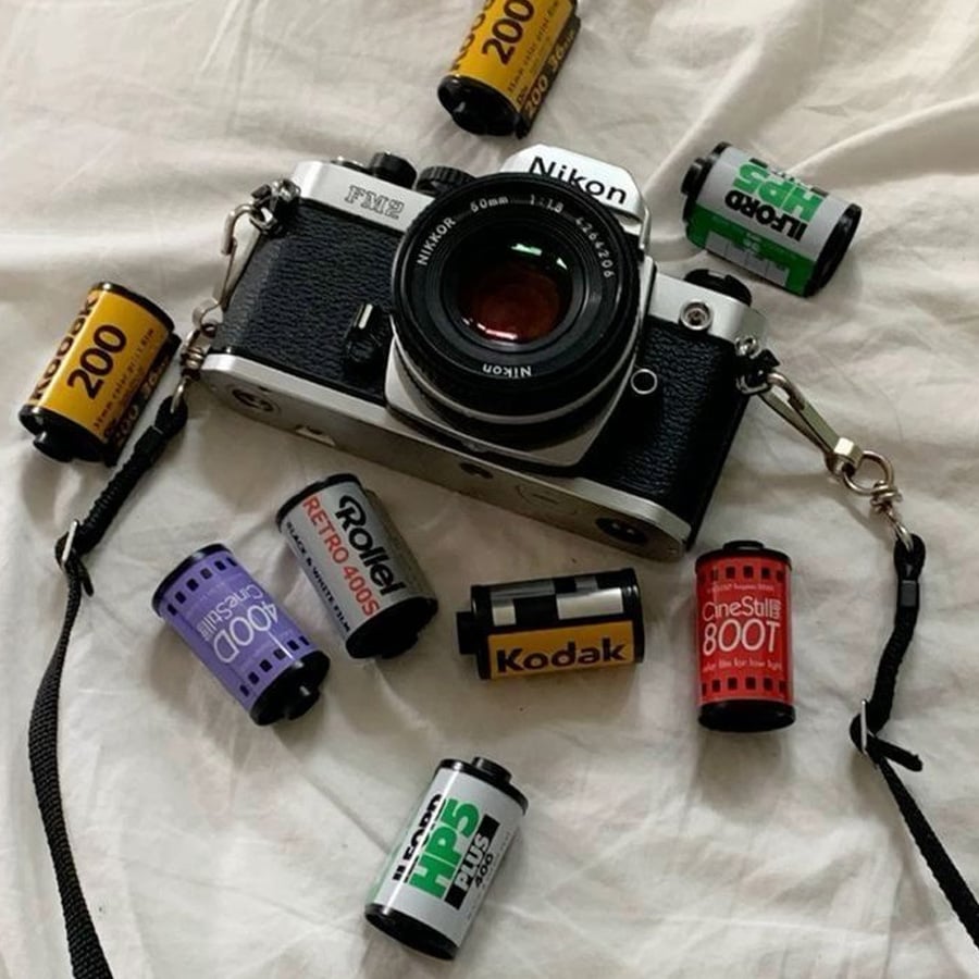 A Nikon film camera lies on a white surface, surrounded by various 35mm film rolls, including Kodak, Ilford, CineStill, and Rollei brands.