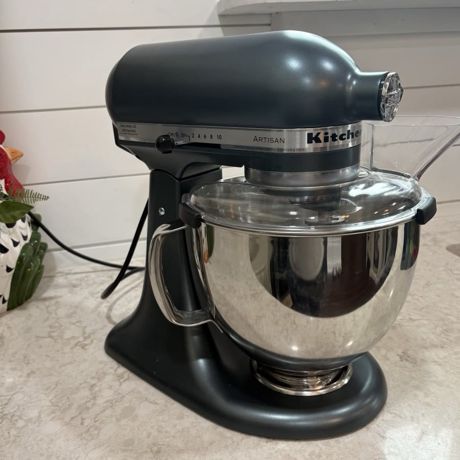 A gray KitchenAid Artisan stand mixer with a stainless steel bowl sits on a kitchen countertop near a white shiplap wall.