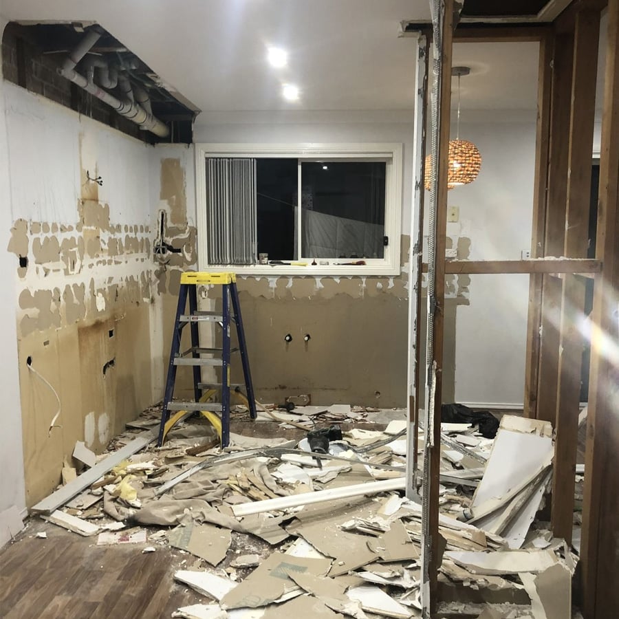 A room under renovation with exposed wall studs, debris on the floor, and a blue stepladder by a window. The walls have missing drywall and visible pipes and fittings.