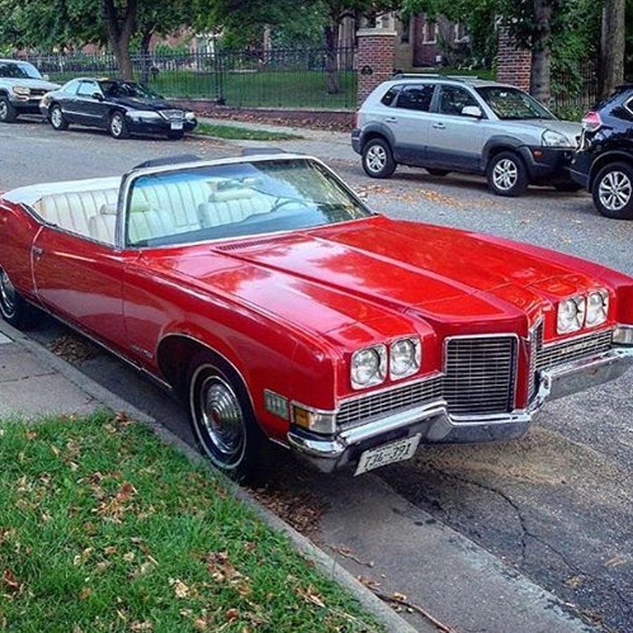 A shiny red vintage convertible car is parked on a residential street, with its white top down and white interior seats visible. Other cars and houses are in the background.