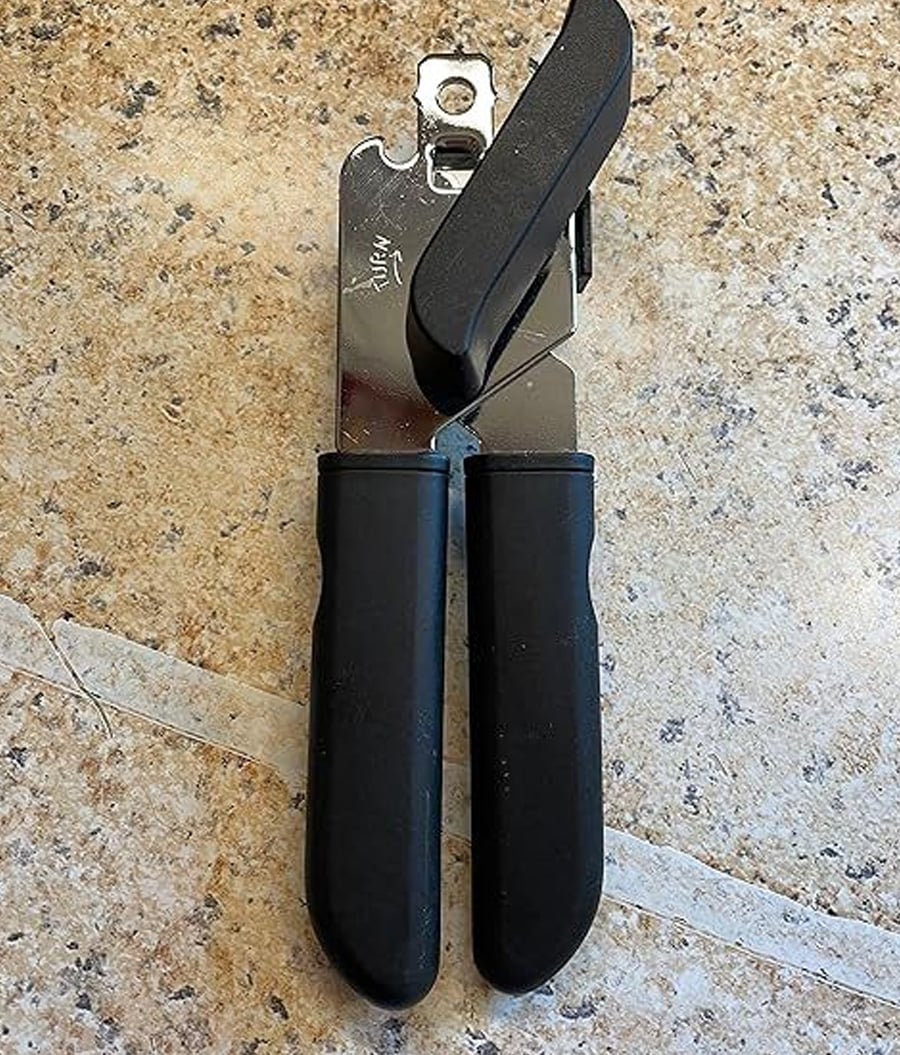 A metal can opener with black handles is lying on a speckled countertop, with its handles closed and the turning knob in view.