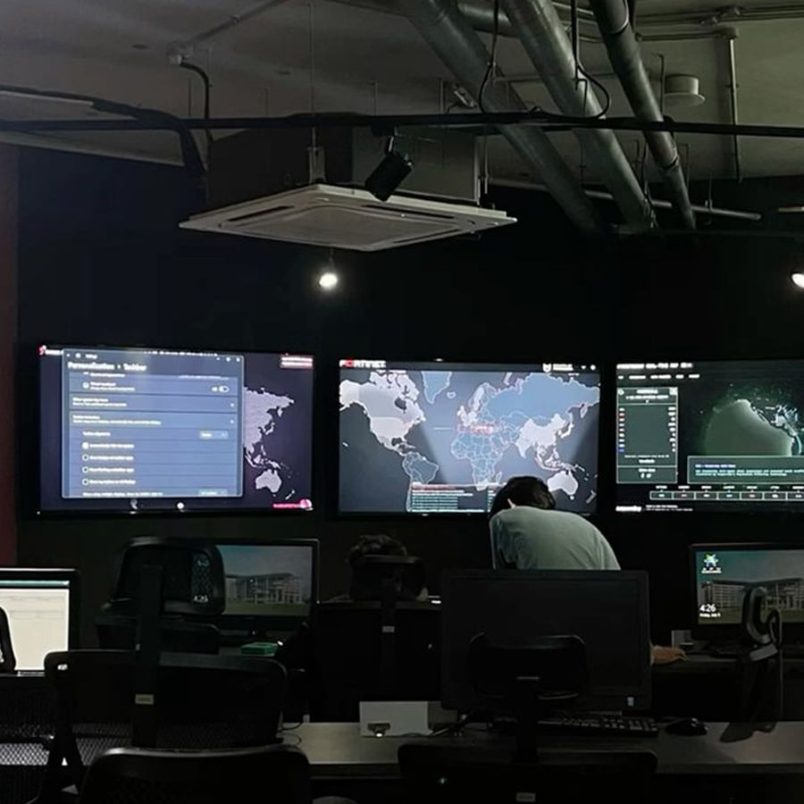 A dimly lit control room with people at desks, large monitors displaying world maps, data, and dashboards on the wall.