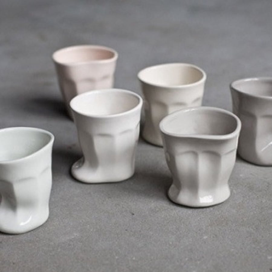 Six uniquely shaped, wavy ceramic cups in soft pastel shades are arranged on a gray surface. Each cup has an irregular, distorted form, giving them an artistic and handmade appearance.